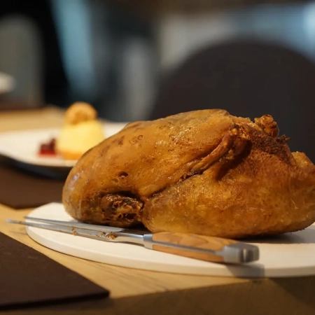 Eine goldbraun gebratene Vierländer Ente auf einem weißen Teller, vor dem unscharfen Hintergrund einer Esstischdekoration im Restaurant Scherrer mit Besteck und Dessertteller. - Ihr Landhaus Scherrer aus Hamburg