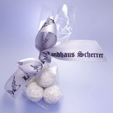 Eine Packung zarter, weiß gepulverter Pralinenselektion-Trüffel, zusammengebunden mit einer eleganten Schleife mit der Aufschrift „Landhaus Scherrer“ und dem sanft bläulichen Hintergrund, der den Eindruck erweckt, es handele sich um eine besondere Köstlichkeit aus einem bestimmten Betrieb. - Ihr Landhaus Scherrer aus Hamburg