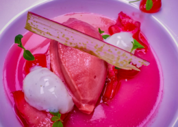 Ein lebendiges Dessert mit Erdbeersorbet, Mousse und Coulis, garniert mit frischen Minzblättern, einer dünnen Waffel und einer ganzen Erdbeere auf einem leuchtend rosa Teller, das einen frischen, fruchtigen Genuss hervorruft. - Ihr Landhaus Scherrer aus Hamburg