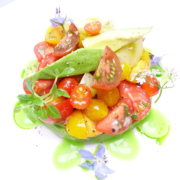 Ein farbenfroher Tomaten-Avocado-Salat, garniert mit essbaren Blüten und frischen Kräutern und beträufelt mit einem würzigen grünen Dressing, bietet eine erfrischende Mischung aus Farben und Aromen. - Ihr Landhaus Scherrer aus Hamburg