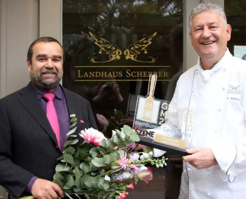 Zwei lächelnde Männer stehen vor dem „Landhaus Scherer“, einer hält einen Blumenstrauß in der Hand, der andere zeigt stolz eine Auszeichnung für herausragende gastronomische Leistungen des Jahres 2010. - Ihr Landhaus Scherrer aus Hamburg