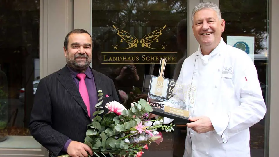 Zwei lächelnde Männer stehen vor dem „Landhaus Scherer“, einer hält einen Blumenstrauß in der Hand, der andere zeigt stolz eine Auszeichnung für herausragende gastronomische Leistungen des Jahres 2010. - Ihr Landhaus Scherrer aus Hamburg