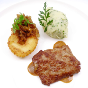 Gegrilltes Steak mit einem Klecks Kräuterkartoffelpüree und einem knusprigen, goldbraunen Kartoffelpuffer, garniert mit karamellisierten Zwiebeln und einem Zweig Petersilie. - Ihr Landhaus Scherrer aus Hamburg