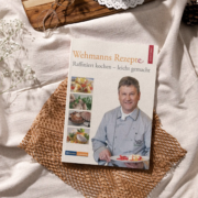 Ein Kochbuch mit dem Titel „Wehmanns Rezepte“ wird auf einem strukturierten Stoff neben kulinarischen Zutaten und Küchengeschirr präsentiert und suggeriert eine Szene der gastronomischen Zubereitung und raffinierten Küche rund um traditionelle deutsche Gerichte - Ihr Landhaus Scherrer aus Hamburg