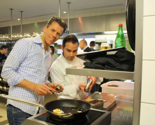 Zwei Köche in einer professionellen Küche, einer lächelt in die Kamera, während er Eier in einer Pfanne kocht, im Hintergrund assistiert ein Koch und Küchengeräte. Sie bereiten Gerichte für einen renommierten nordischen - Ihr Landhaus Scherrer aus Hamburg