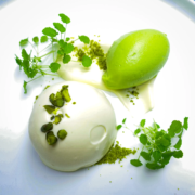 Eine runde Kugel cremeweißen Desserts, garniert mit gehackten Pistazien und begleitet von einer leuchtend grünen Kugel Sorbet, erinnert an die raffinierte Kunstfertigkeit der Hamburger Guide Michelin-Restaurants. Das Dessert auf dem Teller ist mit Microgreens und kleinen grünen Streuseln verziert, alles vor einem weißen Hintergrund. - Ihr Landhaus Scherrer aus Hamburg