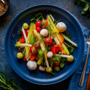 Auf einem tiefblauen Teller wird ein farbenfroher Salat serviert, der an ein exquisites Gericht aus dem Landhaus Scherrer erinnert. Der Salat besteht aus Kirschtomaten, Gurken, Radieschen, Karotten, Grünzeug und Mini-Mozzarella-Kugeln, garniert mit Kräutern. Gabel und Messer liegen auf einer blauen Serviette neben dem Teller, drumherum eine kleine Schüssel mit Gewürzen und frischen Kräutern. - Ihr Landhaus Scherrer aus Hamburg