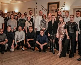 Eine bunt gemischte Gruppe von Menschen, wahrscheinlich das Team vom Landhaus Scherrer, versammelt sich lächelnd und kameradschaftlich unter einem „Frohes neues Jahr!“-Schild für ein Gruppenfoto in Hamburg - Ihr Landhaus Scherrer aus Hamburg