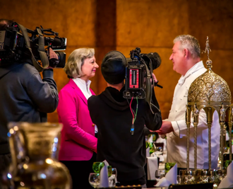 Ein Kameramann filmt ein Interview zwischen einer Frau im rosa Blazer und einem Mann in einer Kochjacke in einem eleganten Ambiente mit reich verziertem Silberbesteck und fängt so einen Blick hinter die Kulissen eines vermutlich gastronomischen Events oder Features ein. - Ihr Landhaus Scherrer aus Hamburg